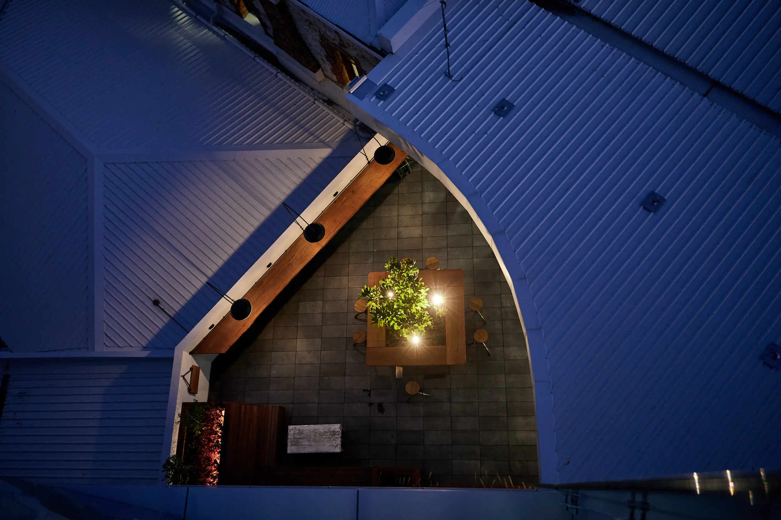 Aerial night view down into the Atwell Arcade courtyard in Fremantle, showing a communal timber table and fig tree lit from below and a row of pendant lights along a timber beam