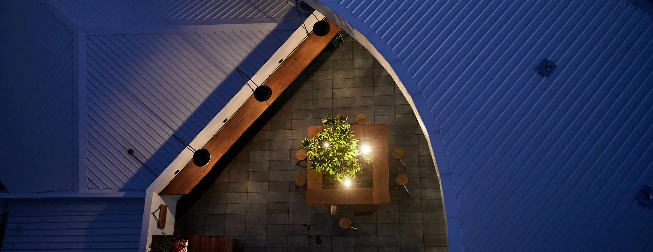 Night view of the Atwell Arcade alfresco courtyard from above, with a lit communal table, feature garden lighting and pendant lights along the beam