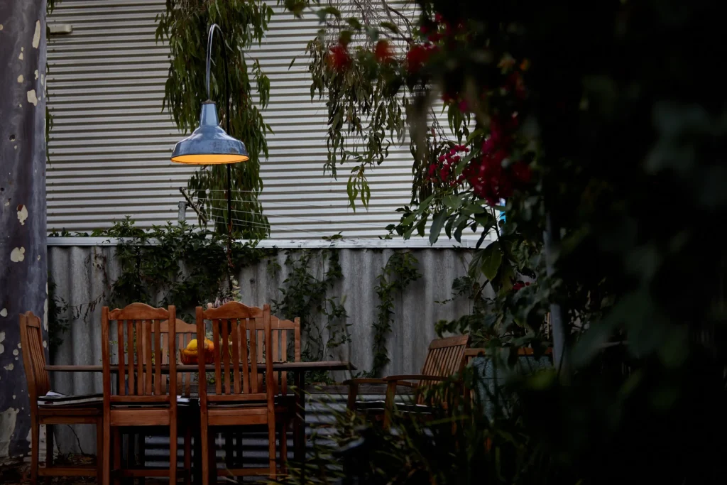 Warehouse-style pendant light hanging above an outdoor dining table with the supporting arm running down to its mounting at the edge of the courtyard