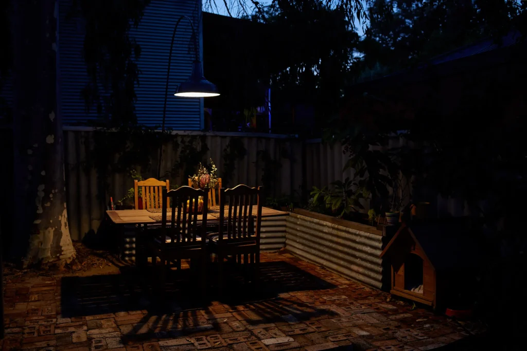 Outdoor dining table and chairs in a Fremantle brick courtyard at night, lit by a single warm pendant light on a curved arm